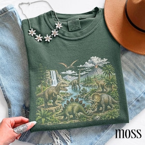 Op de afbeelding: Een mosgroene sweatshirt met een gedetailleerde prehistorische sc&egrave;ne met verschillende dinosaurussen, een waterval en een vulkaan. Het woord "moss" staat rechtsonder gedrukt. Een zilveren ketting met bloemvormige hangers ligt op het shirt.