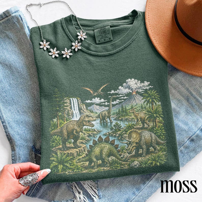Op de afbeelding: Een mosgroene sweatshirt met een gedetailleerde prehistorische sc&egrave;ne met verschillende dinosaurussen, een waterval en een vulkaan. Het woord "moss" staat rechtsonder gedrukt. Een zilveren ketting met bloemvormige hangers ligt op het shirt.