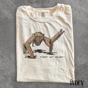 Puede incluir: Camiseta de color marfil con un gráfico de una criatura marrón, parecida a la madera, con detalles verdes, en pose. El texto "FIST MY BUMP!" está impreso debajo. La camiseta está doblada y tiene una etiqueta Comfort Colors.