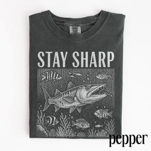 May include: Dark grey t-shirt with the words "STAY SHARP" above a detailed illustration of a barracuda and other fish in a marine environment. The shirt is a Comfort Colors brand.