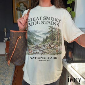May include: Ivory-colored t-shirt featuring a scenic illustration of the Great Smoky Mountains National Park. The design includes a mountain landscape, trees, a river, and deer. The text "Great Smoky Mountains National Park, Tennessee, USA" is printed on the shirt.