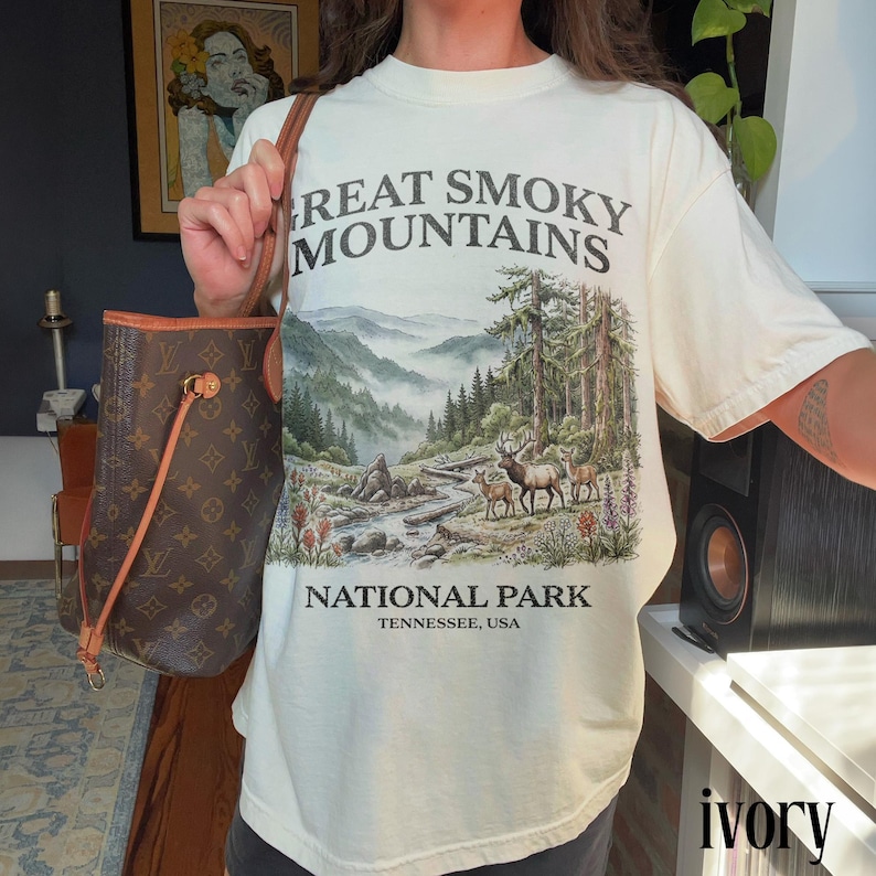 May include: Ivory-colored t-shirt featuring a scenic illustration of the Great Smoky Mountains National Park. The design includes a mountain landscape, trees, a river, and deer. The text "Great Smoky Mountains National Park, Tennessee, USA" is printed on the shirt.
