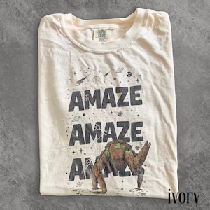 May include: Ivory-colored t-shirt with the word "AMAZE" printed three times in dark gray, along with a graphic of a space creature. The shirt also features stars, planets, and comets.