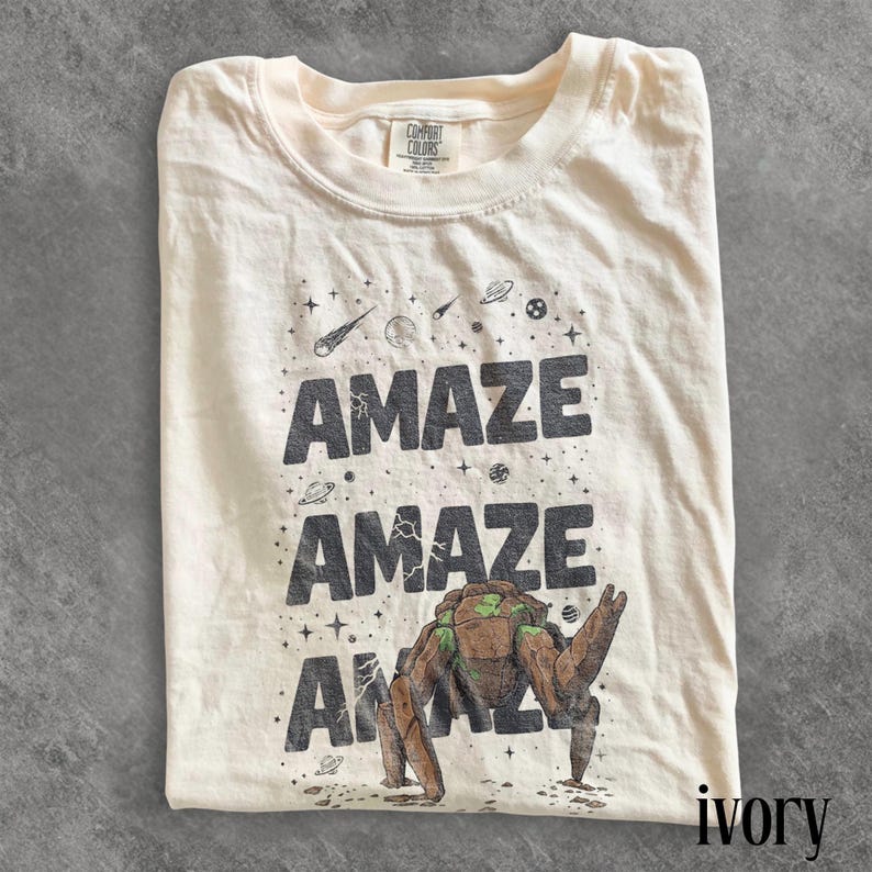May include: Ivory-colored t-shirt with the word "AMAZE" printed three times in dark gray, along with a graphic of a space creature. The shirt also features stars, planets, and comets.