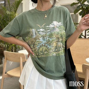 Op de afbeelding: Olijfgroen T-shirt met een gedetailleerd dinosauruslandschap ontwerp. De afbeelding bevat verschillende dinosaurussen, een waterval en een vulkaan. Het shirt heeft korte mouwen en een relaxte pasvorm.