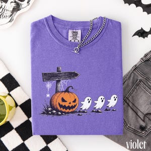 May include: A purple short-sleeve t-shirt featuring a graphic of a jack-o'-lantern, a wooden signpost, and three small white ghosts. The shirt has a silver-toned chain necklace draped around the collar and a Comfort Colors brand label.