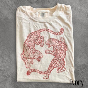 May include: Ivory-colored t-shirt featuring a red illustration of two leopards in a dynamic pose. The shirt has a crew neck and short sleeves. The brand name "Comfort Colors" is visible on the tag. The word "ivory" is printed at the bottom right.