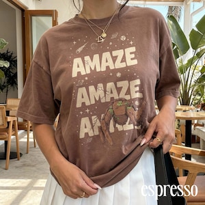 May include: Brown t-shirt with the word "AMAZE" repeated three times in white, along with a graphic of a creature. The shirt has short sleeves and a relaxed fit. The word "espresso" is printed in white on the bottom right.