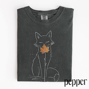 Puede incluir: Una camiseta de cuello redondo gris carbón con una ilustración de líneas blancas de un zorro que sostiene una hoja de arce naranja en la boca. La camiseta tiene una etiqueta Comfort Colors en el cuello y la palabra pepper impresa en negro.