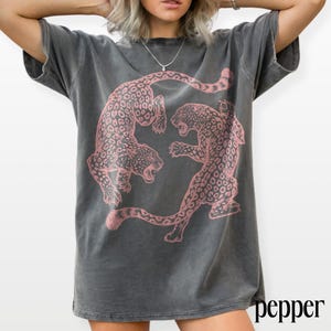 May include: Dark gray t-shirt featuring a pink leopard print design. The graphic shows two leopards facing each other. The word "pepper" is printed in black on the bottom right corner of the shirt.