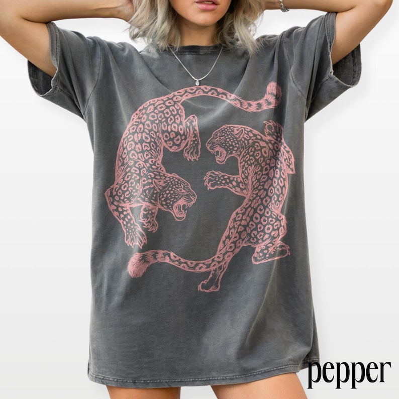 May include: Dark gray t-shirt featuring a pink leopard print design. The graphic shows two leopards facing each other. The word "pepper" is printed in black on the bottom right corner of the shirt.