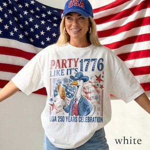 May include: White t-shirt with a patriotic design featuring a goose in a suit holding a corn dog, with the text "PARTY LIKE IT'S 1776" and "USA 250 YEARS CELEBRATION". The design incorporates red, white, and blue colors.