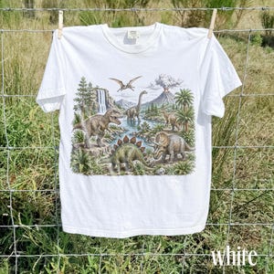 Op de afbeelding: Wit t-shirt met een gedetailleerde illustratie van dinosaurussen in een prehistorisch landschap. Het ontwerp bevat een waterval, vulkaan en palmbomen. Het woord "white" staat onderaan het shirt.