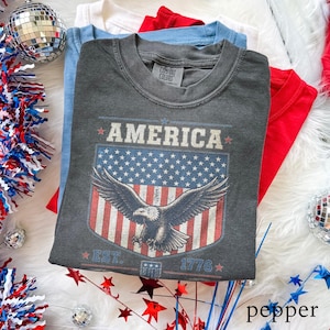 May include: Folded t-shirts in red, white, and blue. The top shirt is pepper grey with an American flag design featuring an eagle and the word "AMERICA". The design includes the text "EST. 1776".
