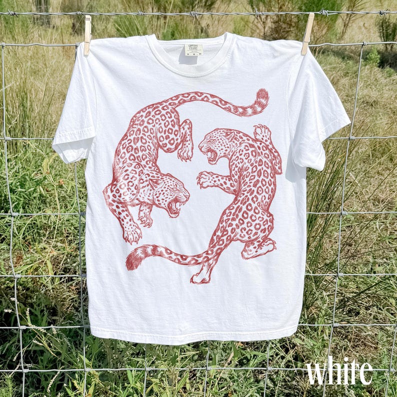 May include: White t-shirt featuring a red illustration of two leopards in a dynamic pose. The leopards have spotted patterns. The shirt is hanging on a clothesline. The word "white" is at the bottom of the image.