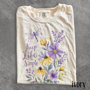 May include: An ivory t-shirt featuring a watercolor-style graphic of purple and yellow wildflowers, lavender sprigs, and a dragonfly. The text "Keep Life Simple" is printed in a purple script font on the left side of the floral arrangement.