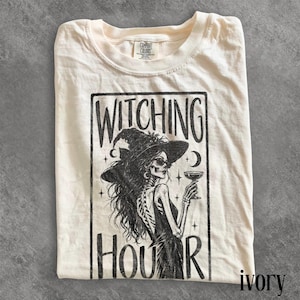 May include: An ivory t-shirt featuring a black graphic print of a skeleton wearing a witch hat and sunglasses while holding a cocktail glass. The text WITCHING HOUR is printed in a bold font above and below the illustration within a rectangular frame.