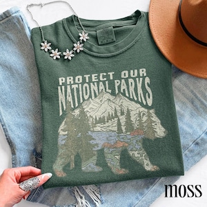 May include: A moss green t-shirt with the text "PROTECT OUR NATIONAL PARKS" and a graphic of a bear with a mountain and forest scene. A silver necklace with flower accents and a brown hat are also visible.