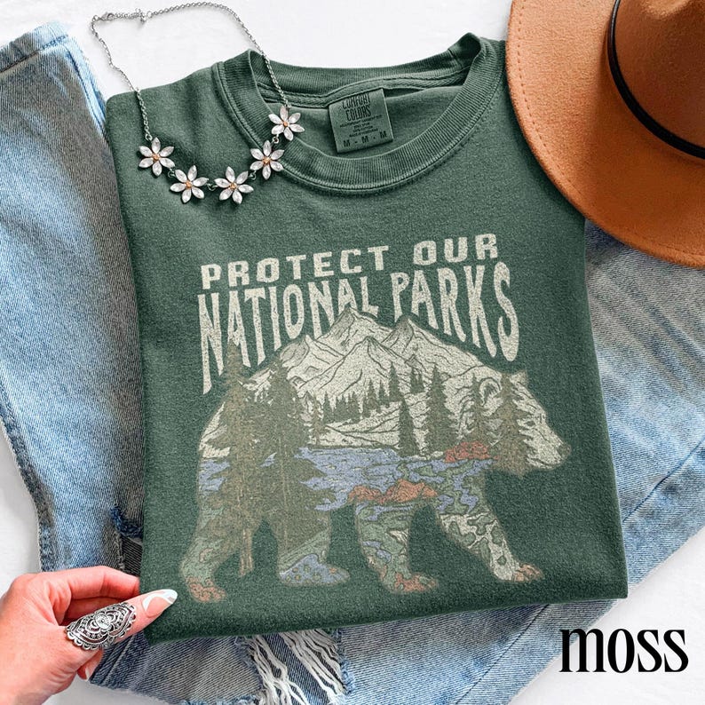 May include: A moss green t-shirt with the text "PROTECT OUR NATIONAL PARKS" and a graphic of a bear with a mountain and forest scene. A silver necklace with flower accents and a brown hat are also visible.
