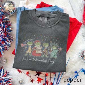 Könnte beinhalten: Ein dunkelgraues Comfort Colors T-Shirt mit einer Grafik von Cartoon-Fröschen in patriotischer Kleidung, mit dem Text "Just an Independent Frog" und "Since 1776". Das Shirt ist mit roten, weißen und blauen Shirts gestapelt.