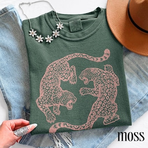 May include: Moss green sweatshirt featuring a pink leopard print design. The sweatshirt has a crew neck and long sleeves. A silver necklace with flower-shaped pendants is draped on the shirt. The word "moss" is printed on the bottom right.