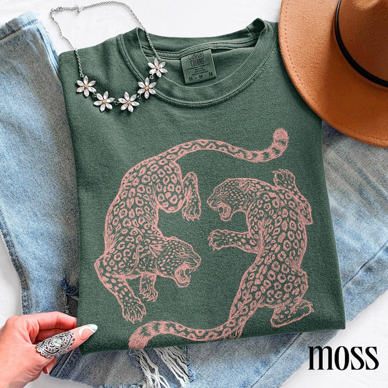May include: Moss green sweatshirt featuring a pink leopard print design. The sweatshirt has a crew neck and long sleeves. A silver necklace with flower-shaped pendants is draped on the shirt. The word "moss" is printed on the bottom right.