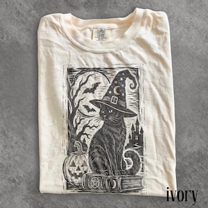 May include: An ivory t-shirt featuring a black ink-style graphic of a black cat wearing a witch hat, sitting on a book. The scene includes a jack-o'-lantern, a full moon, bats, and a castle silhouette. The inner neck label reads Comfort Colors.