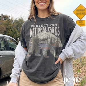 May include: Dark gray t-shirt with the text "PROTECT OUR NATIONAL PARKS" above a graphic of a bear, mountains, and trees. The shirt is worn with a gray zip-up hoodie and tan pants.