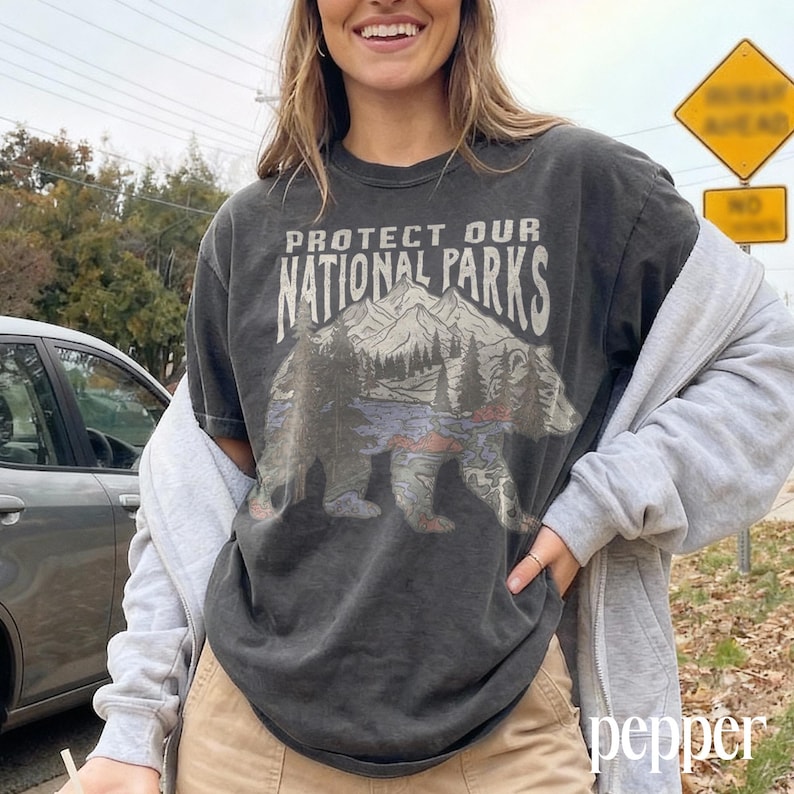 May include: Dark gray t-shirt with the text "PROTECT OUR NATIONAL PARKS" above a graphic of a bear, mountains, and trees. The shirt is worn with a gray zip-up hoodie and tan pants.
