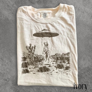Könnte beinhalten: Elfenbeinfarbenes T-Shirt mit einer Grafik eines Hundes, der von einem UFO entführt wird. Der Hund steht auf seinen Hinterbeinen und blickt auf das Raumschiff. Das Shirt hat ein Wüstenlandschaftsdesign.