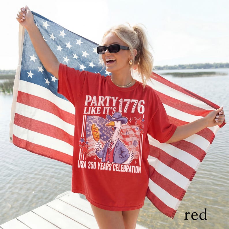 May include: Red t-shirt with the text "PARTY LIKE IT'S 1776" and "USA 250 YEARS CELEBRATION." The graphic features a goose in a suit holding a corn dog. The person is holding an American flag.