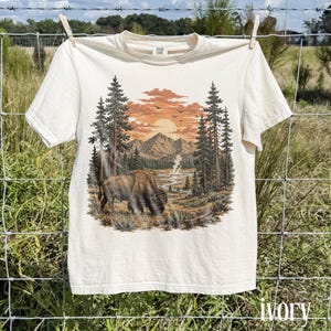 May include: An ivory short-sleeve t-shirt featuring a graphic print of a brown bison grazing in a landscape with tall pine trees, a winding river, distant mountains, and a sunset sky with birds flying above the horizon in the background.