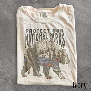 May include: Ivory-colored t-shirt with the text "PROTECT OUR NATIONAL PARKS" above a graphic of a bear with a mountain and forest scene. The shirt is folded and has a Comfort Colors tag.