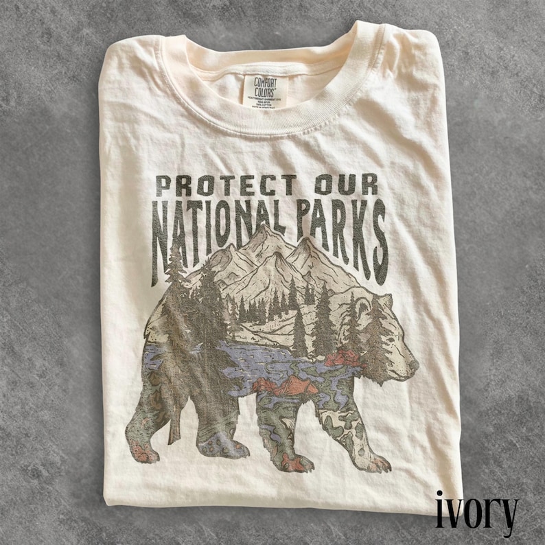 May include: Ivory-colored t-shirt with the text "PROTECT OUR NATIONAL PARKS" above a graphic of a bear with a mountain and forest scene. The shirt is folded and has a Comfort Colors tag.