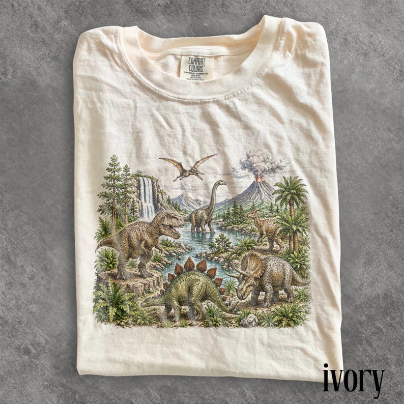 Op de afbeelding: Ivoor kleurig T-shirt met een gedetailleerde prehistorische sc&egrave;ne met verschillende dinosaurussen, een vulkaan en een waterval. Het shirt heeft een ronde hals en het merk "Comfort Colors" is zichtbaar. Het woord "ivory" staat rechtsonder.