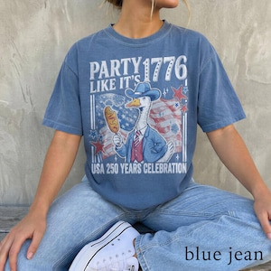 May include: Blue t-shirt with a graphic of a goose wearing a cowboy hat and suit, holding a corn dog. The shirt has the text "PARTY 1776 LIKE IT'S" and "USA 250 YEARS CELEBRATION". The person is wearing blue jeans and white sneakers.