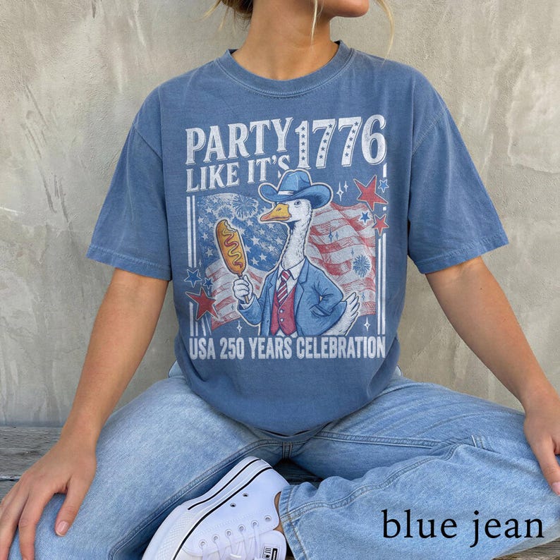 May include: Blue t-shirt with a graphic of a goose wearing a cowboy hat and suit, holding a corn dog. The shirt has the text "PARTY 1776 LIKE IT'S" and "USA 250 YEARS CELEBRATION". The person is wearing blue jeans and white sneakers.