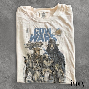 May include: Ivory-colored t-shirt featuring a graphic of cow characters in a parody of a space opera. The text "COW WARS" is printed in blue above the characters. The shirt is made of a soft material.