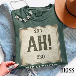 May include: A moss green t-shirt featuring a distressed graphic with the text "AH!" and numbers. A silver necklace with flower pendants and a brown hat are also visible. The graphic includes the text "THE ELEMENT" at the bottom.