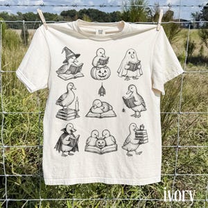 May include: An off-white short-sleeve t-shirt featuring a black ink graphic print of nine distinct duck illustrations. The ducks are depicted in various scenes, including wearing a witch hat, dressed as a ghost, reading books, and sitting on pumpkins.
