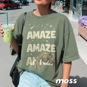 May include: Moss green t-shirt with the word "AMAZE" repeated three times, with a space-themed graphic. The word "moss" is printed at the bottom right. The shirt is worn by a person.