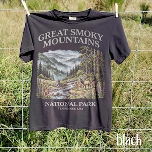 May include: Black t-shirt featuring a scenic illustration of the Great Smoky Mountains National Park. The design includes a river, trees, mountains, and deer. Text on the shirt reads "Great Smoky Mountains National Park, Tennessee, USA."