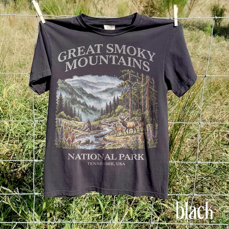 May include: Black t-shirt featuring a scenic illustration of the Great Smoky Mountains National Park. The design includes a river, trees, mountains, and deer. Text on the shirt reads "Great Smoky Mountains National Park, Tennessee, USA."
