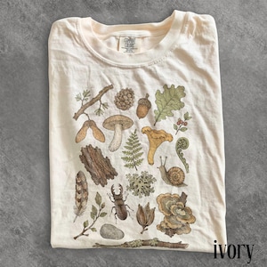 May include: An ivory t-shirt featuring a detailed botanical and woodland illustration. The graphic includes a brown beetle, a snail, a feather, various mushrooms, an acorn, a pinecone, tree bark, a fern, an oak leaf, and twigs.