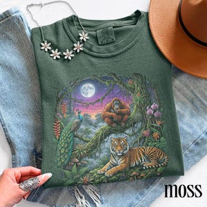 Puede incluir: Sudadera verde de cuello redondo con una escena de la jungla que presenta un tigre, un orangután y un pavo real. El diseño incluye una luna llena y colores vibrantes. La palabra "moss" está impresa en la parte inferior derecha.