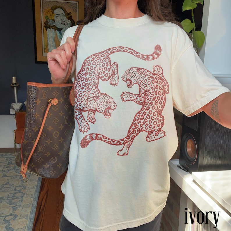 May include: Ivory-colored t-shirt featuring a red illustration of two leopards in a dynamic pose. The shirt has a classic crew neck and short sleeves. The word "ivory" is printed at the bottom right corner of the image.