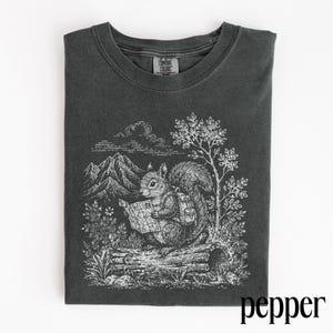 May include: A charcoal gray t-shirt featuring a white line art graphic of a squirrel wearing a backpack and holding a map while sitting on a log. The background includes mountains, trees, and foliage. The inner neck label reads Comfort Colors.