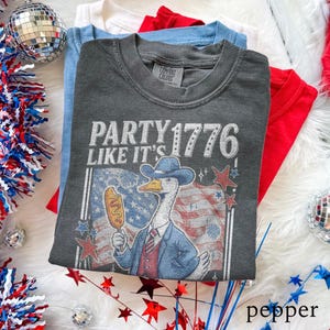May include: A stack of folded t-shirts in red, white, and blue. The top shirt is dark gray with the text "PARTY 1776 LIKE IT'S" and a cartoon goose wearing a cowboy hat and holding a corn dog. The shirts are on a white, fluffy surface.