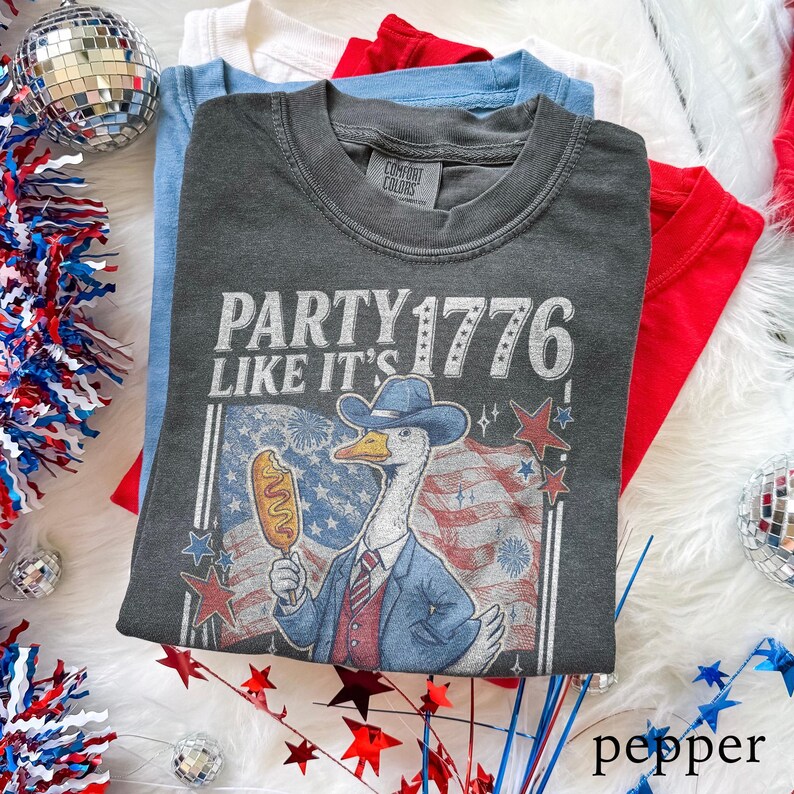 May include: A stack of folded t-shirts in red, white, and blue. The top shirt is dark gray with the text "PARTY 1776 LIKE IT'S" and a cartoon goose wearing a cowboy hat and holding a corn dog. The shirts are on a white, fluffy surface.