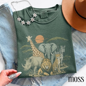 May include: A dark green t-shirt featuring a graphic print of a giraffe, elephant, zebra, rhinoceros, lion, and cheetah under a sun. The shirt has a crew neckline and a Comfort Colors brand label. A silver flower-shaped necklace is placed on top.